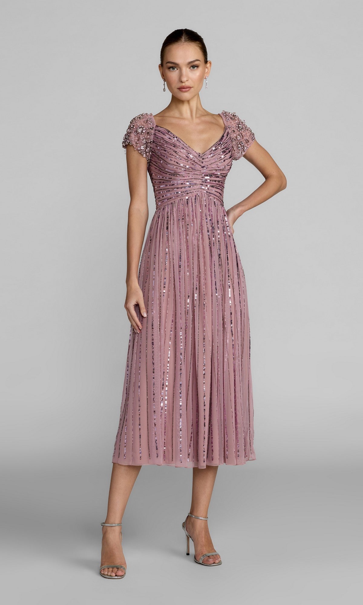 A woman in the Mac Duggal 23098 midi party dress in vintage lilac, featuring beaded short sleeves, a ruched bodice, and sequin embellishments, paired with silver high-heeled sandals, stands against a plain gray background.